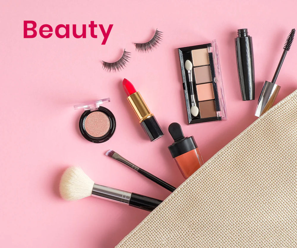 Beauty and makeup items arranged on a pink background, including false eyelashes, a lipstick, an eyeshadow palette, a mascara tube, a liquid blush bottle, brushes, and a textured beige cosmetic pouch. The word 'Beauty' appears in pink text in the upper left.